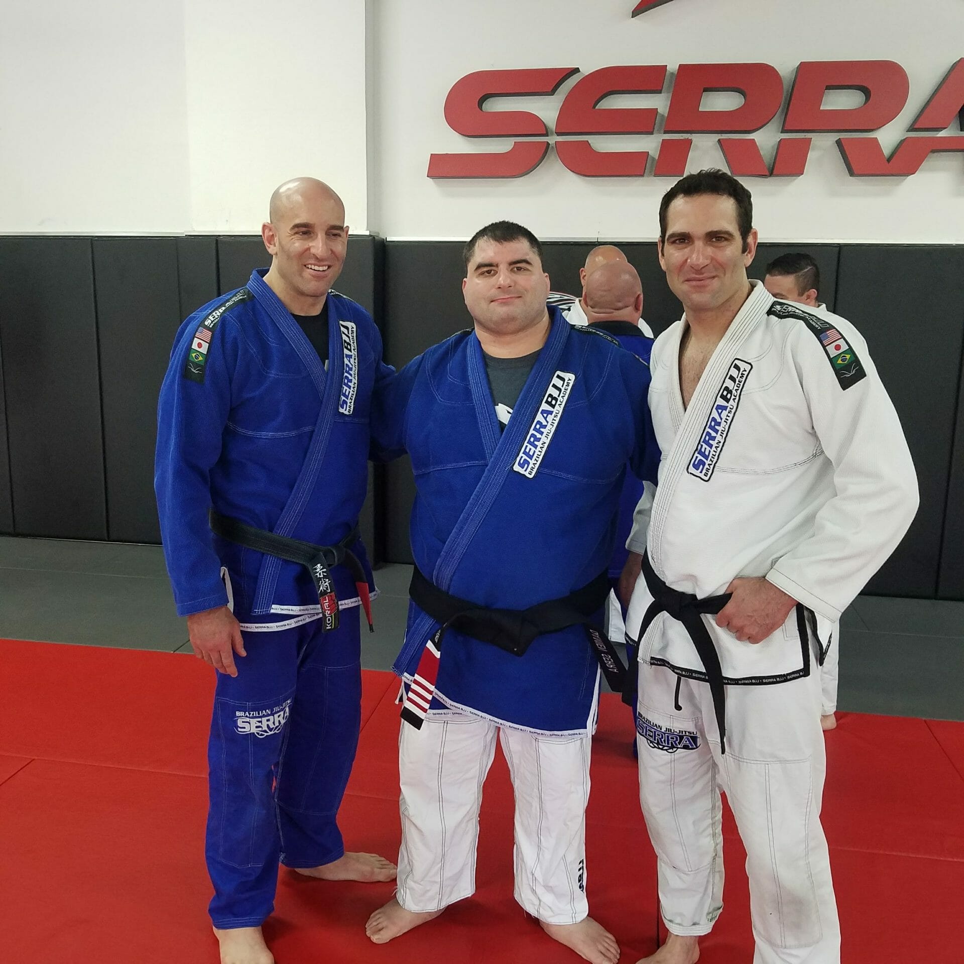 Israel Joffe in a white jiu-jitsu gi with training partners in front of the Serra Jiu-Jitsu sign